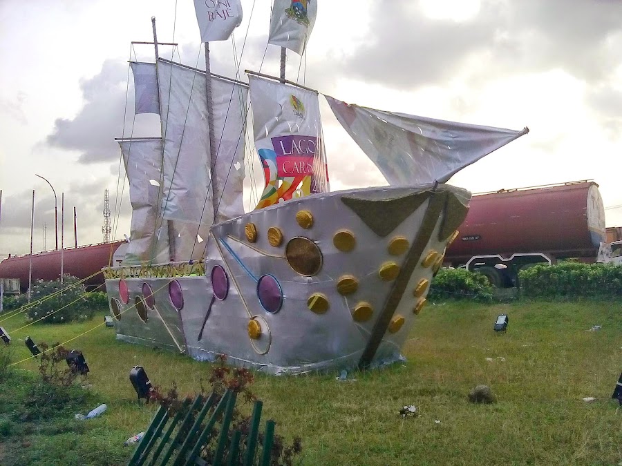 The White Ship, The ship raised as a roadside display, its sails and flags printed ‘Lagos Carnival’ and ‘Eko Oni Baje’; tanker wagons behind.